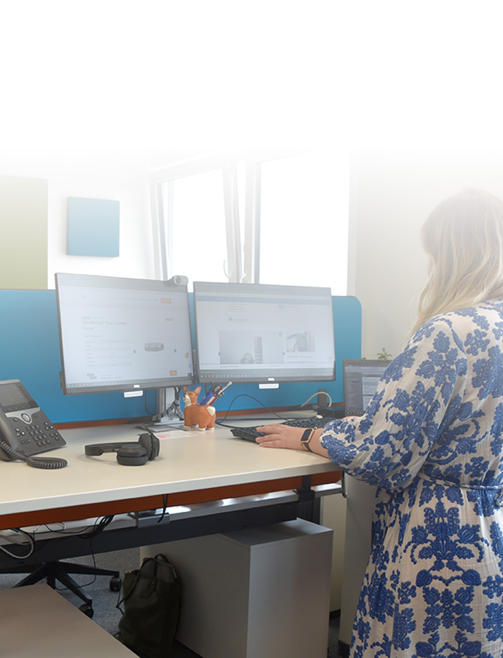 A woman stands at a desk with two monitors.